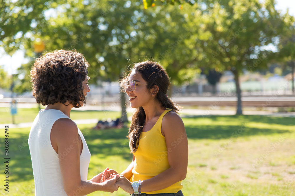 Fototapeta premium Happy young women talking in park. Cheerful multiethnic female friends greeting each other and holding hands in park. Friendship concept