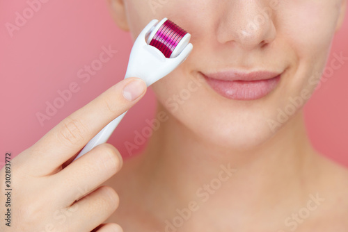 Close-up female face with dermaroller for mesotherapy procedures, skin care at home and in salon. Meso roller with microneedles on pink background photo