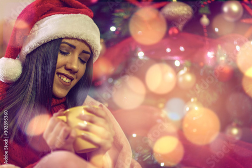 Smiling girl in Santa hat drinking tea over christmas treeat home photo