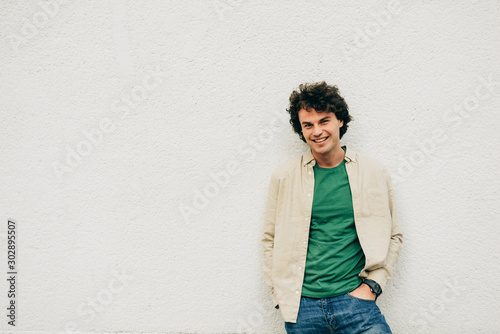 Image of cheerful young man posing for advertisement, smiling and looking at the camera, standing at building concrete wall outdoors. Portrait of happy student male with curly hair resting outside.