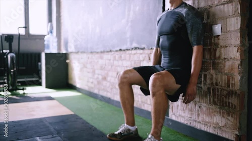 Male fitness instructor does wall sit exercise at gym. Wall sits to make hip and thigh stronger. Effective exercises for running. Short flexibility-focus session at workout place.