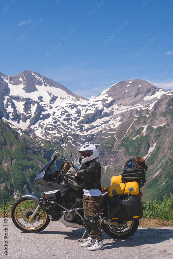 Woman in full biker outfit, jacket protection turtle. Touring motorcycle with big bags. The snowy peaks of the Alpine mountains on background. Grossglockner Pass. vertical photo. Austria, Italy