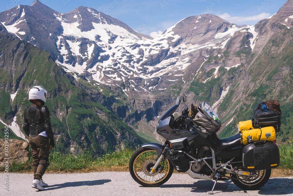 Woman in full biker outfit, jacket protection turtle. Touring motorcycle with big bags. The snowy peaks of the Alpine mountains on background. Grossglockner Pass Hochalpenstrasse. Austria, Italy