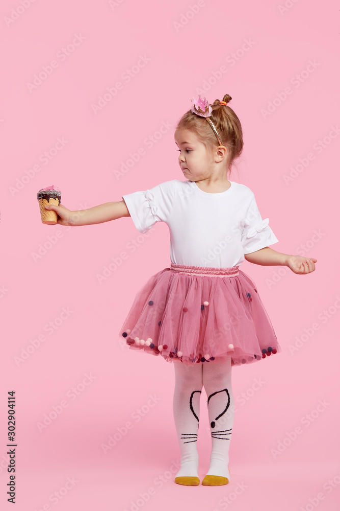 Cute child girl holds ice cream on his outstretched arm and covers his mouth with his hand. Refuses to eat a tasteless dessert on a pink studio background. Proper baby food without sugar