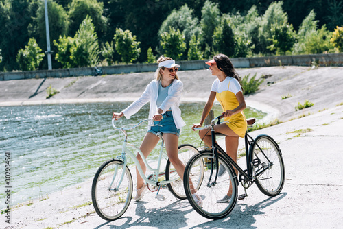 happy blonde and brunette girls with bikes looking at each other near river in summer