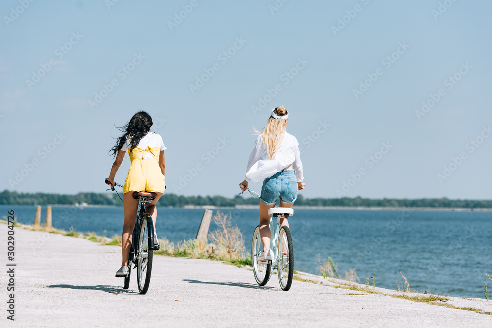 back view of blonde and brunette girls riding bikes near river in summer