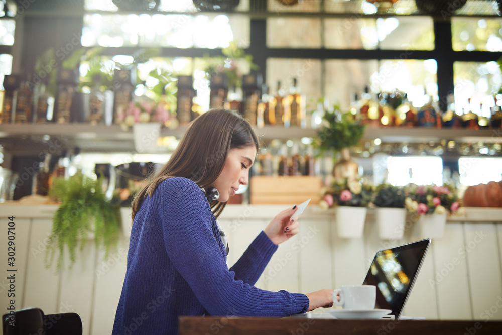Beautiful elegant caucasian brunette in sweater sitting in cafe and using laptop for online shopping. In hand is credit card.