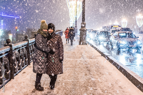 Two girls in warm winter clothes in heavy snow are standing on the illuminated sidewalk. Nearby are driving cars.