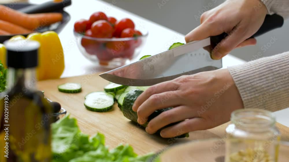 healthy eating, vegetarian food and cooking concept - young woman with kitchen knife chopping cucumber on wooden cutting board at home