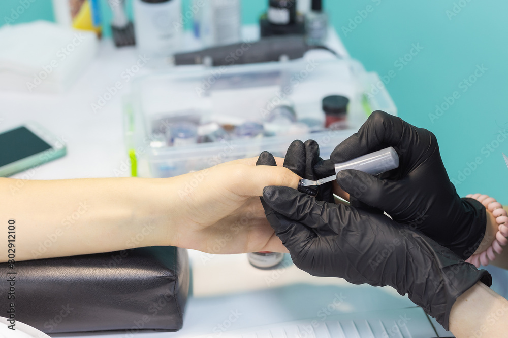 Closeup top view of female hands of professional manicurist in black gloves holding hands of client and applying transparent finish top polish at black manicure at nail. Horiozntal color photography. Closeup top view of female hands of professional manicurist in black gloves holding hands of client and applying transparent finish top polish at black manicure at nail. Horiozntal color photography.