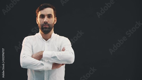 Young man in white shirt, standing on the black background, posing, folding his hands, looking confidently at the camera.