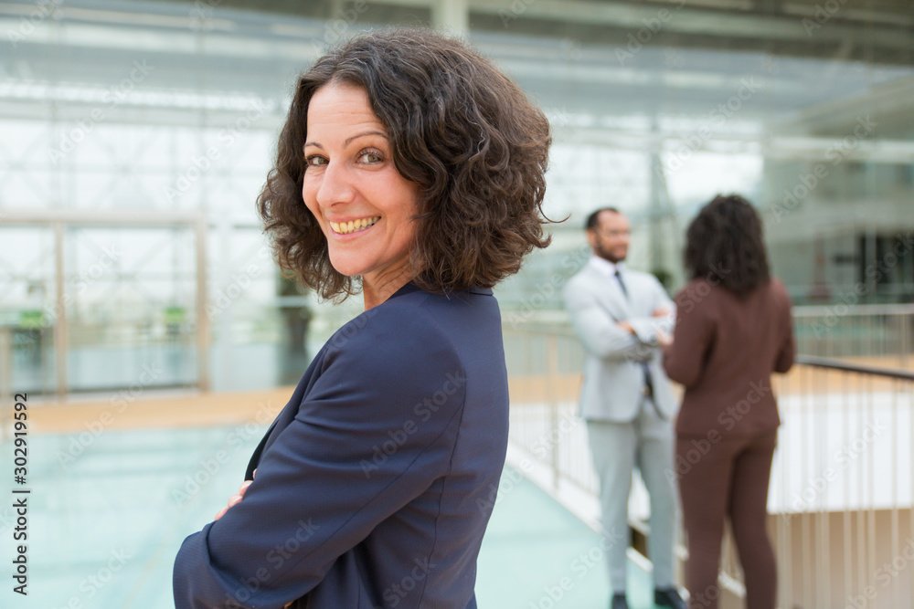 Obraz premium Happy successful business woman posing with arms folded, looking at camera, smiling. Her male and female colleagues standing and talking in background. Confident business leader concept