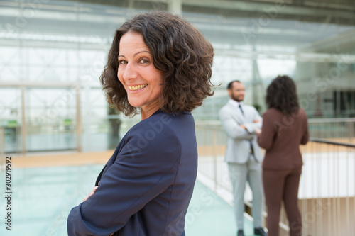Free Download Happy successful business woman posing with arms folded, looking at camera, smiling. Her male and female colleagues standing and talking in background. Confident business leader concept - GFXLoad