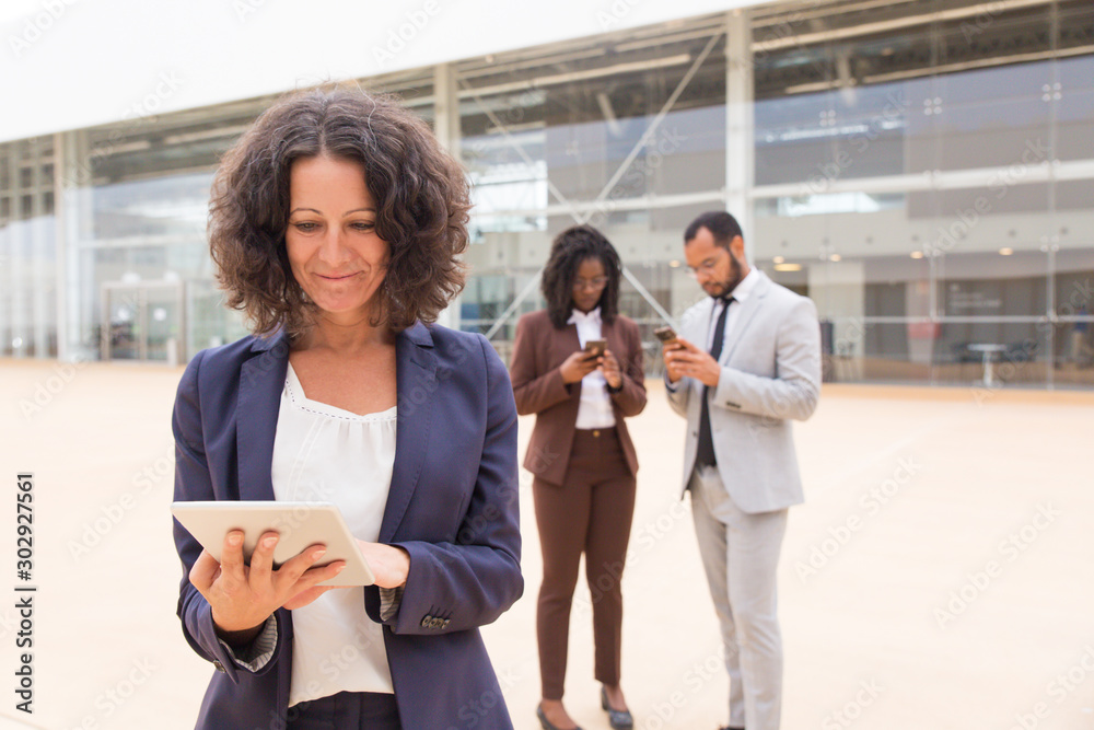 Obraz premium Happy satisfied business woman using tablet outside office building. Her male and female colleagues using mobile phones in background. Wireless internet connection concept