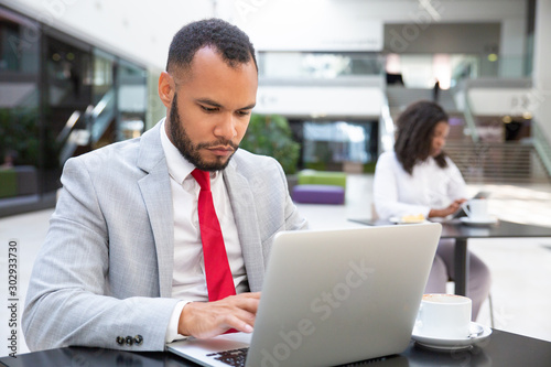 Focused serious professional using laptop while drinking coffee in office lobby. Young African American woman using tablet in background. Wireless technology concept