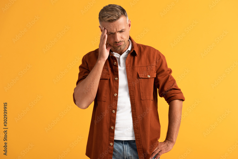 exhausted man touching temple while standing with hand on hip isolated on orange exhausted man touching temple while standing with hand on hip isolated on orange