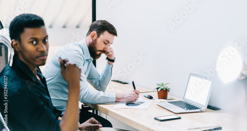 Concentrated man writing information of idea for startup project while african colleague on blurred frontage looking at camera, two male entrepreneurs collaborating in company office during work time