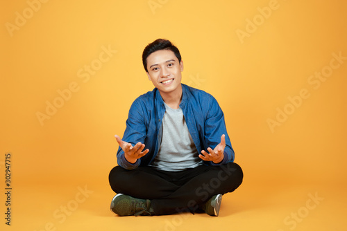 Good looking young Asian man sitting with legs crossed on the floor, isolated on orange background. © makistock