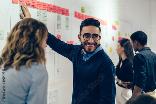 Positive young man laughing while collaborating with colleagues on creating presentation using colorful stickers for productive work in office.Male and female students having fun during workshop photo