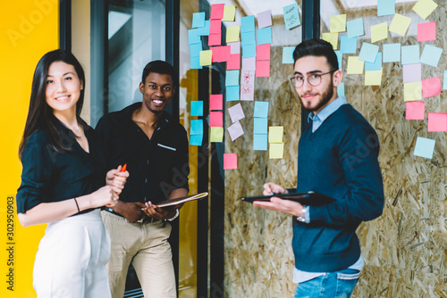 Portrait of multicultural team of skilled students smiling at camera while standing near wall with colorful stickers for learning new words during courses.Diverse group of young creative designers photo