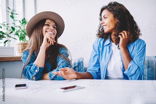 Cheerful female best friends in stylish casual wear having fun on meeting in cafe interior resting together, happy hipster girls communicating and joking while sitting in coffee shop during free time