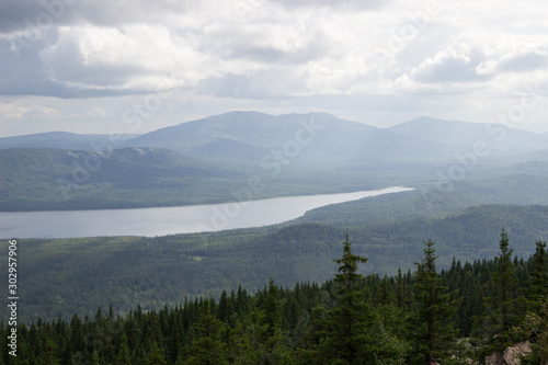 Beautiful mountain landscape with a lake in the middle of a spruce forest on a cloudy day, a photo from a height in the foreground bright spruce trees with fading colors as they move away
