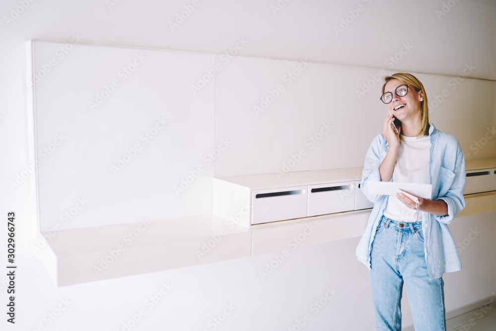 Cheerful young woman in eyewear talking with friend on smartphone while holding envelope with letter after checking mailbox.Positive hipster girl laughing during mobile conversation in hallway Cheerful young woman in eyewear talking with friend on smartphone while holding envelope with letter after checking mailbox.Positive hipster girl laughing during mobile conversation in hallway