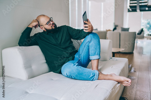 Pensive caucasian male in spectacles sitting on cozy sofa at partment watching video on portable pc, hipster guy in eyewear reading ebook via digital tablet using wireless connection at home photo