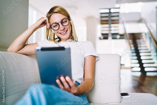 Positive blonde young woman in optical spectacles watching funny tutorial on touch pad resting on couch at home.Cheerful blogger in earphones laughing during reading comments from followers on tablet