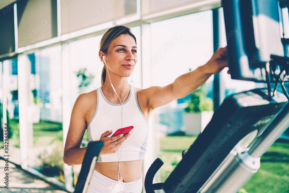 Attractive positive blonde woman dressed in sportswear configuring elliptical cross trainer exercises bike for better training in gym keeping fitness body enjoying music via earphones on smartphone