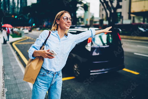 Foto Pretty hipster girl in eyewear raising hand standing on road waving for take a c