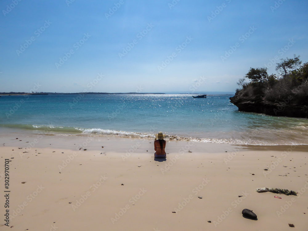 A girl in bikini and straw hat sitting on Pink Beach, Lombok Indonesia. Girl is enjoying the heavenly view. The water has many shades of blue. The beach is surrounded by small hills. Paradise beach.