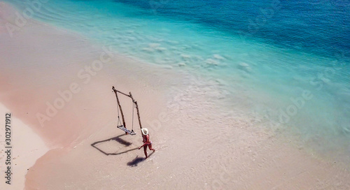 A girl swinging on a swing placed on the seashore of Pink Beach, Lombok, Indonesia. The swing has simple wood construction. Waves wash the pillars of it. In the back there are few boats. Drone capture