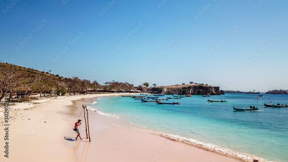 A man swinging on a swing placed on the seashore of Pink Beach, Lombok, Indonesia. The swing has simple wood construction. Waves wash the pillars of it. In the back there are few boats. Drone capture