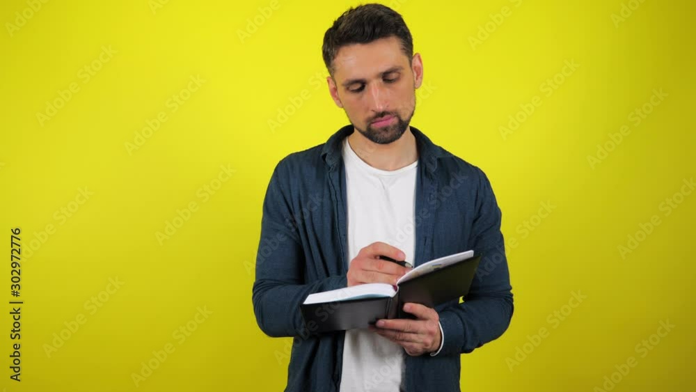 Young man in blue shirt and white T-shirt writes tasks, goals, dreams, ideas in a notebook with a black pen. Yellow background with copy space. 4k slowmotion footage. Young man in blue shirt and white T-shirt writes tasks, goals, dreams, ideas in a notebook with a black pen. Yellow background with copy space. 4k slowmotion footage.