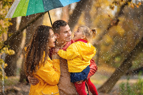 Happy parents and daughter with rainbow colored umbrella under rain on nature