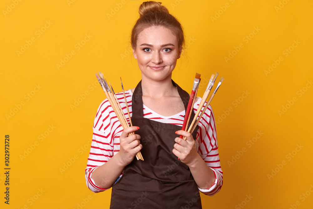 Portrait of positive magnetic young painter posing over yellow background in studio, holding bunch of brushes in both hands, having pleasant facial expression, smiling. Hobby and art concept. Portrait of positive magnetic young painter posing over yellow background in studio, holding bunch of brushes in both hands, having pleasant facial expression, smiling. Hobby and art concept.