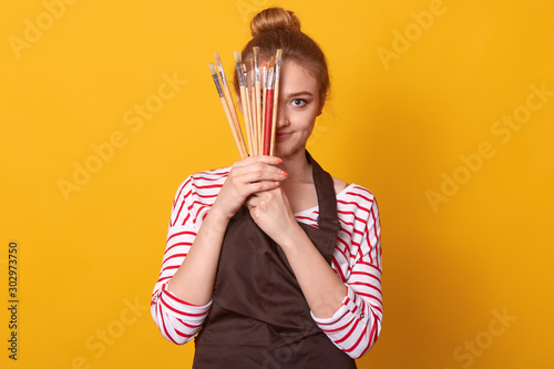 Picture of delighted tender cute young lady holding lots of brushes in both hands, covering half of face with art equipment, wearing striped sweatshirt and brown apron. People and free time concept.