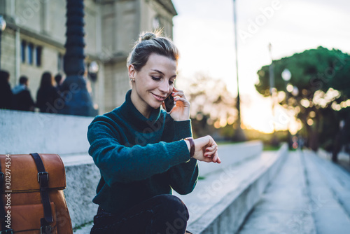 Smiling young woman having phone call while checking wristwatch on street stairs photo
