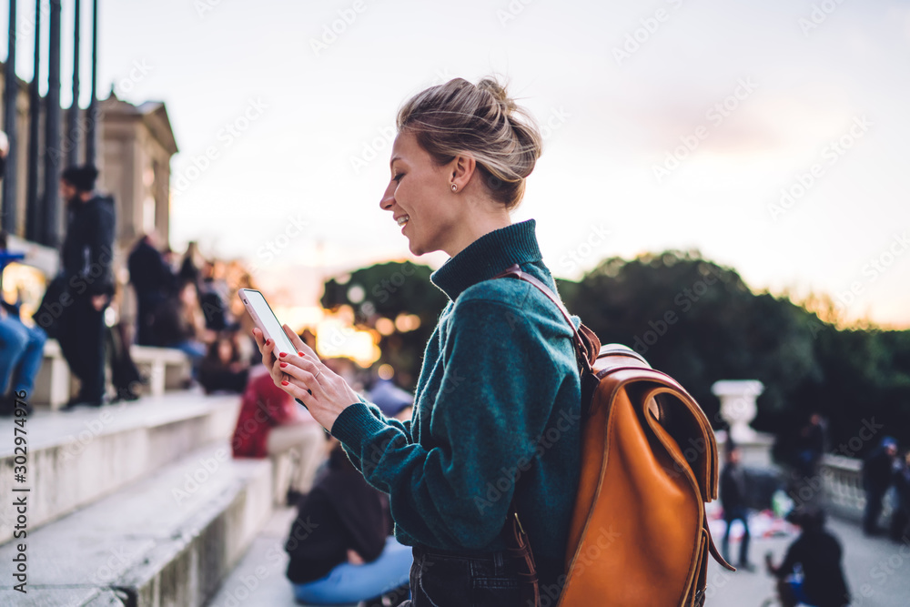 Side view of millennial female tourist in casual apparel chatting at online mobile platform for travelers, happy successful woman reading received email during phoning via modern cellphone gadget - obrazy, fototapety, plakaty 