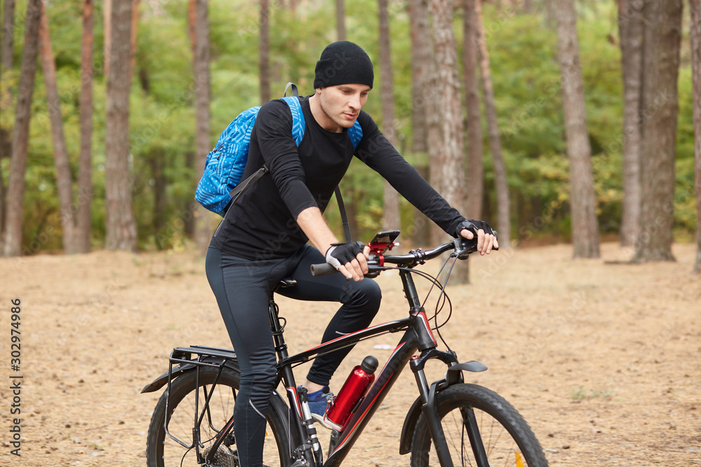 Confident handsome cyclist riding his bike in autumn forest, following his way, having map on his smartphone, wearing black sportswear, having blue backpack, spending time around nature alone. Confident handsome cyclist riding his bike in autumn forest, following his way, having map on his smartphone, wearing black sportswear, having blue backpack, spending time around nature alone.