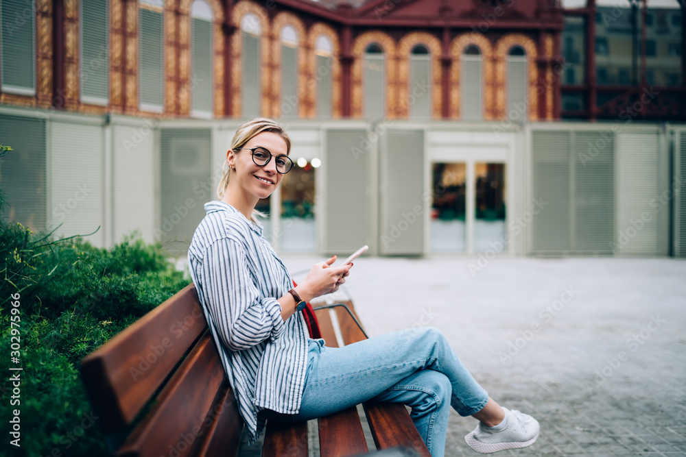 Smiling stylish woman using phone on bench and looking at camera