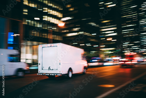 White truck of logistic company driving on city road delivering goods for order quickly in time, truck moving speedly on avenue transporting and distributing stuff for postal service in megalopolis