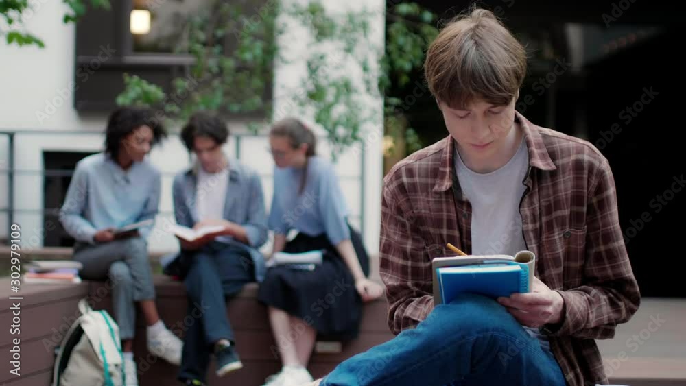 Medium shot of casual student boy thoughtfully studying alone with group of students on background in university campus outdoor
