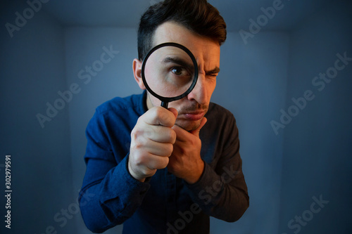 Surprised Young man student holding magnifying glass looking to camera with a pensive emotion isolated over grey  background. Science and curiosity concept.