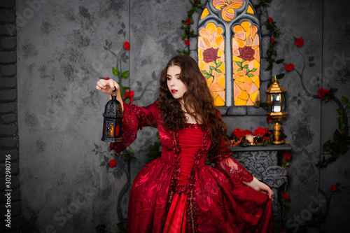 A beautiful girl in a magnificent red dress of the Rococo era stands against a fireplace, a window and flowers with a lamp with candles in her hands.