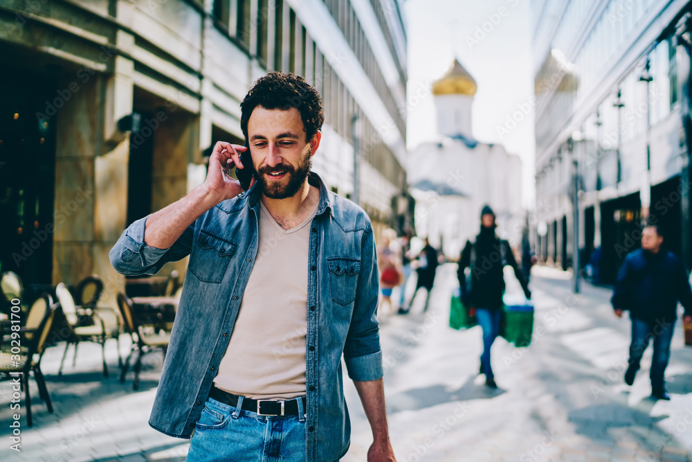 Bearded hipster guy going to shopping in city and calling on smartphone device to talk with friend.Stylish young man dressed in denim wear communicating on mobile phone while walking near mall