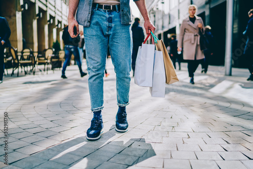 Cropped view of stylish hipster guy customer dressed in stylish denim clothes and fashionable footwear holding paper bags in hands after good shopping during sales in mall standing outdoor on pavement