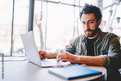 Portrait of bearded it professional looking at camera while sitting at laptop computer and working on freelance on development of design website in modern office using free wireless internet