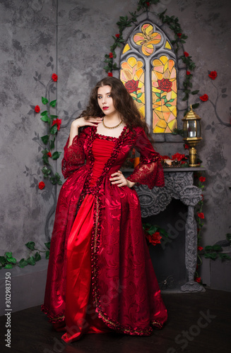 Beautiful girl in a magnificent  red rococo dress. Against the background of the fireplace  window  and flowers.